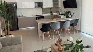 A modern kitchen with white cabinets, a central island with bar stools, a flat-screen TV mounted on the wall, and a dining area with a grey sofa and a coffee table.