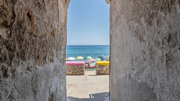 Vista sulla spiaggia attraverso un arco in un muro di pietra, con ombrelloni colorati e cielo sereno.