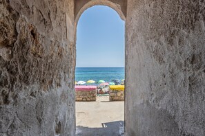 Vista da praia por uma porta em arco com parede de pedra, guarda-sóis coloridos e céu claro.