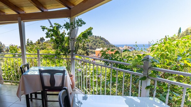 Ein überdachter Balkon mit Tisch und Stühlen, mit Blick auf eine malerische Landschaft mit Bäumen und Meerblick.