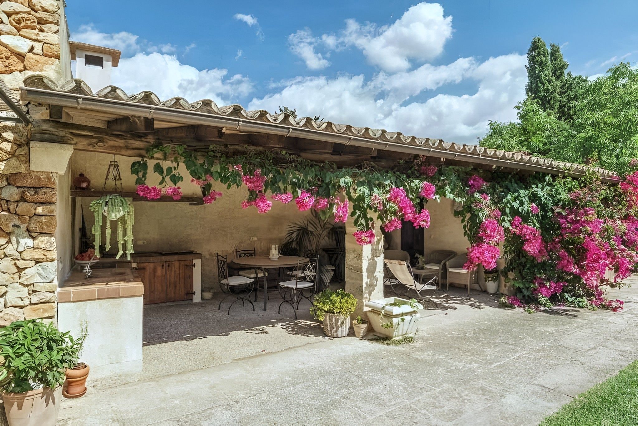A covered outdoor patio with a stone wall, a table and chairs, and flowering plants.
