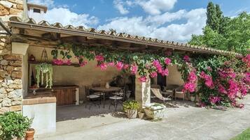 A covered outdoor patio with a stone wall, a table and chairs, and flowering plants.