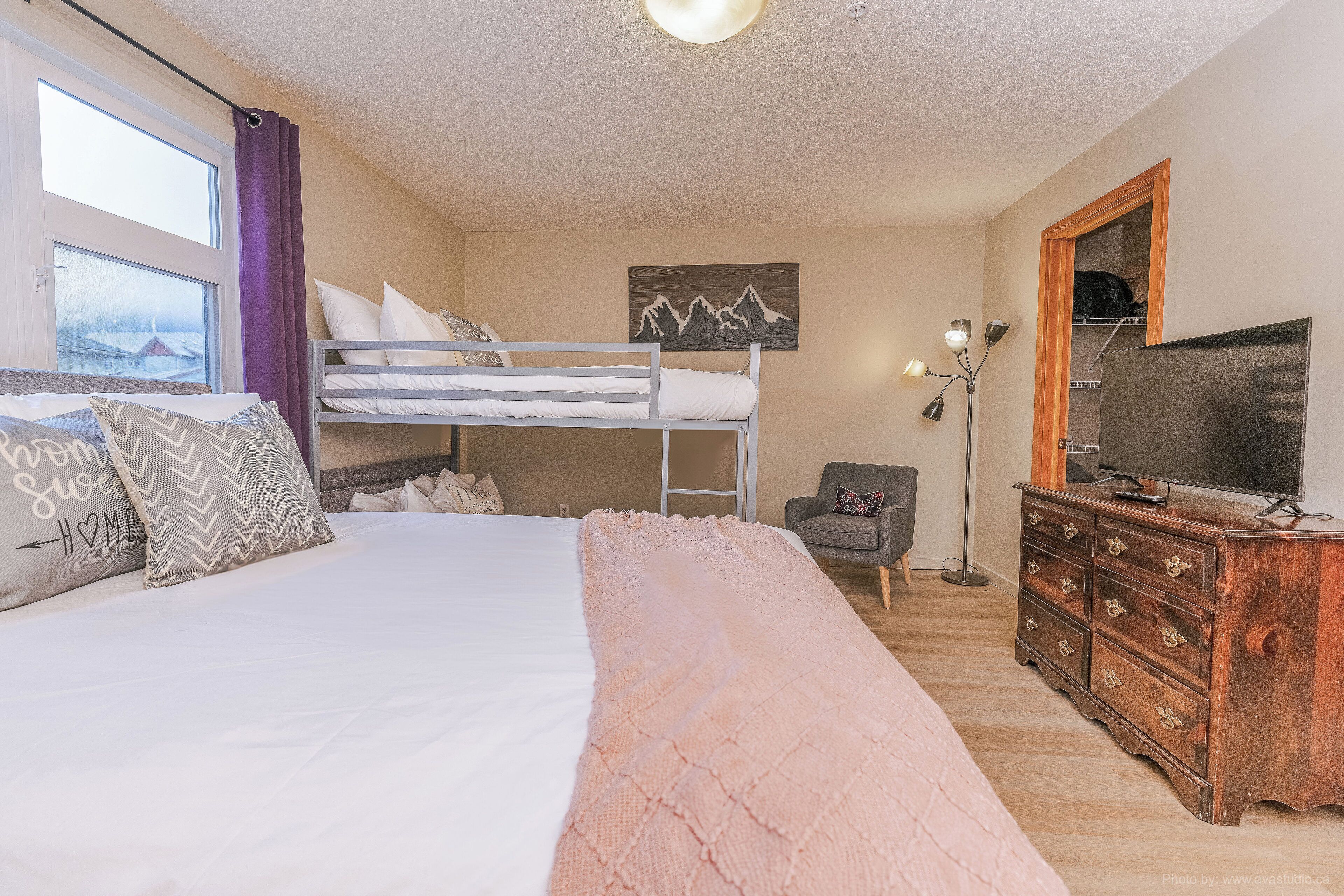 A bedroom with a bunk bed, a desk, a chair, a dresser, a TV, and a window with curtains.