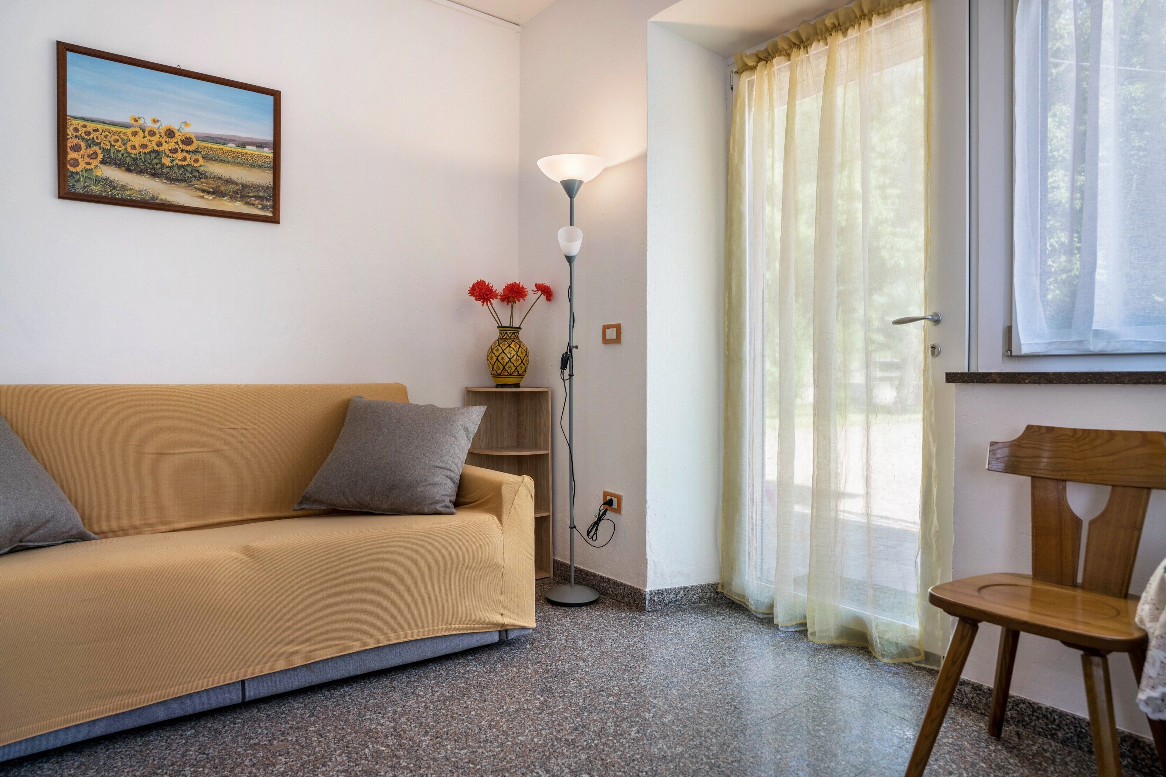 A room with a sofa, a floor lamp, a chair, a vase with flowers, and a painting on the wall.