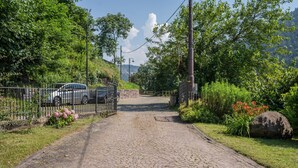 A cobblestone path leading to a gated entrance with a parked car, surrounded by greenery and a clear sky.