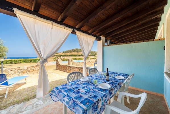 Un área de comedor al aire libre techada, con una mesa dispuesta para dos personas, una piscina al fondo y vistas al paisaje.