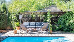 A pool with a wooden deck, a cushioned outdoor seating area, and lush greenery in the background. - Villa 'Es Tancat' with Sea View, Private Pool and Wi-Fi (Sa Ràpita)