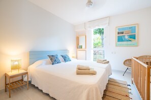 A bedroom with a bed, a nightstand, a crib, a window, and a wall-mounted picture. - Villa 'Es Tancat' with Sea View, Private Pool and Wi-Fi (Sa Ràpita)