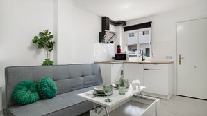 Un espace de vie moderne avec un canapé gris, une petite table à manger et un coin cuisine doté de meubles blancs et d’une hotte noire.