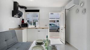 Une cuisine compacte avec un coin repas, un canapé gris et une vue sur la rue par une porte ouverte.