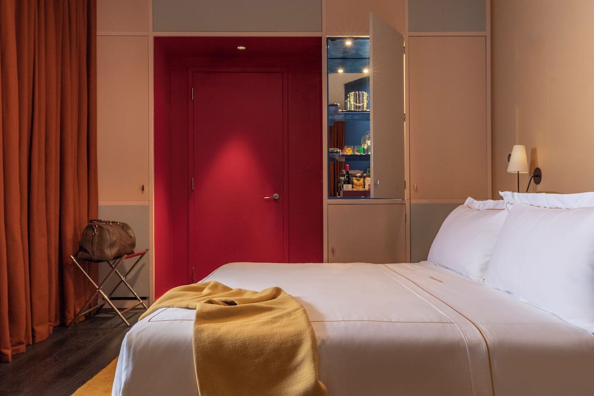 A hotel room with a bed, a red door, a small seating area, and a minibar.