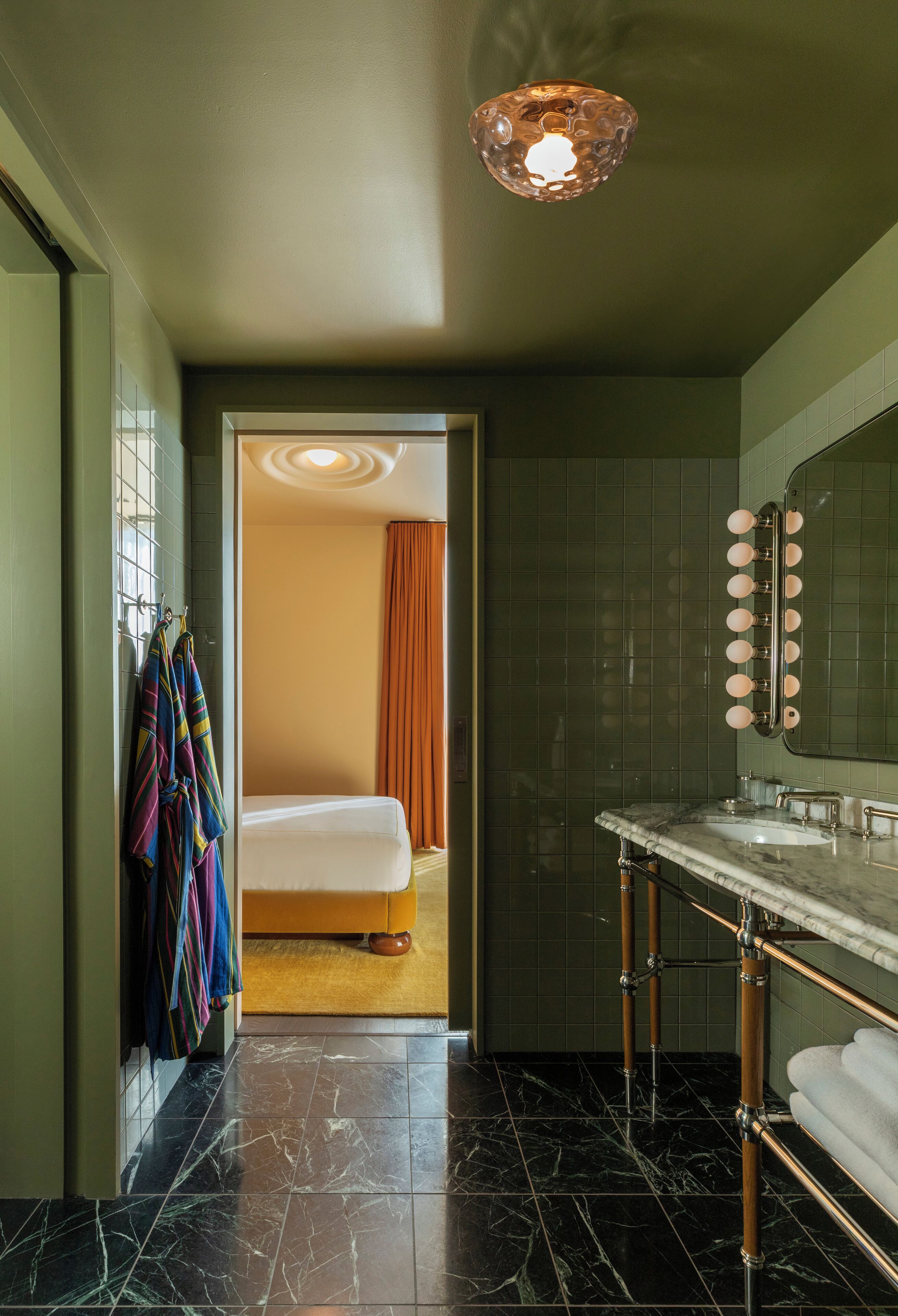Een hotelbadkamer met een marmeren vloer, een wastafel met spiegel, en een bed dat door een open deur zichtbaar is.