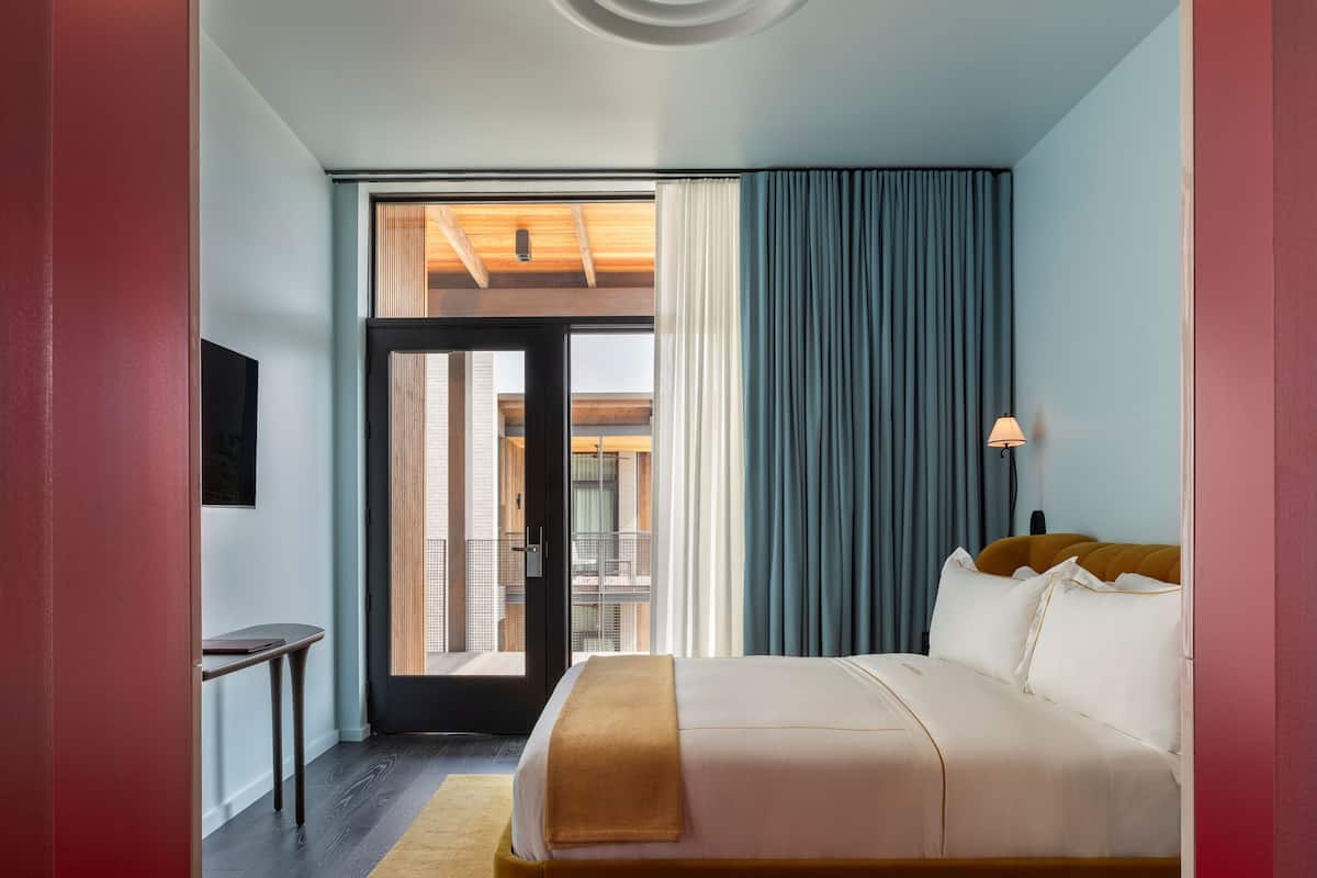 A hotel room with a bed, a desk, a chair, a window with curtains, and a view of the outside.