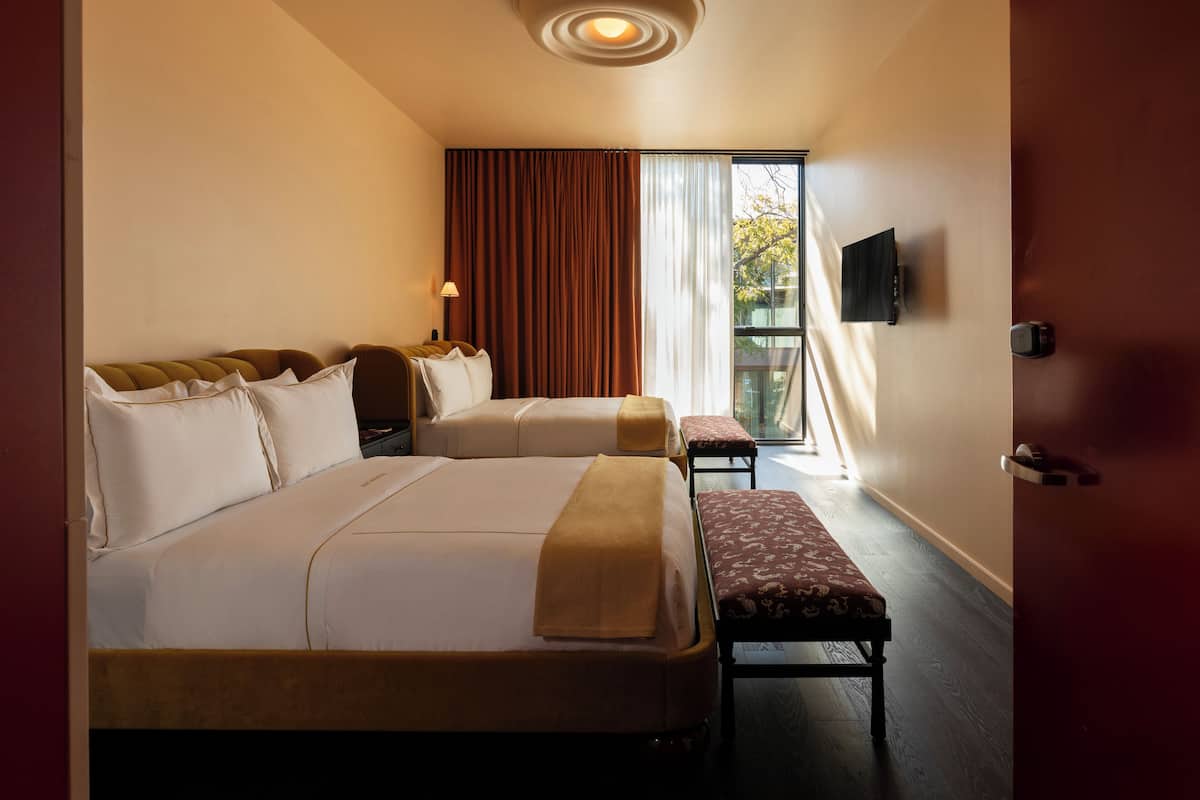 A hotel room with two beds, a bench, a TV, and a window with curtains.