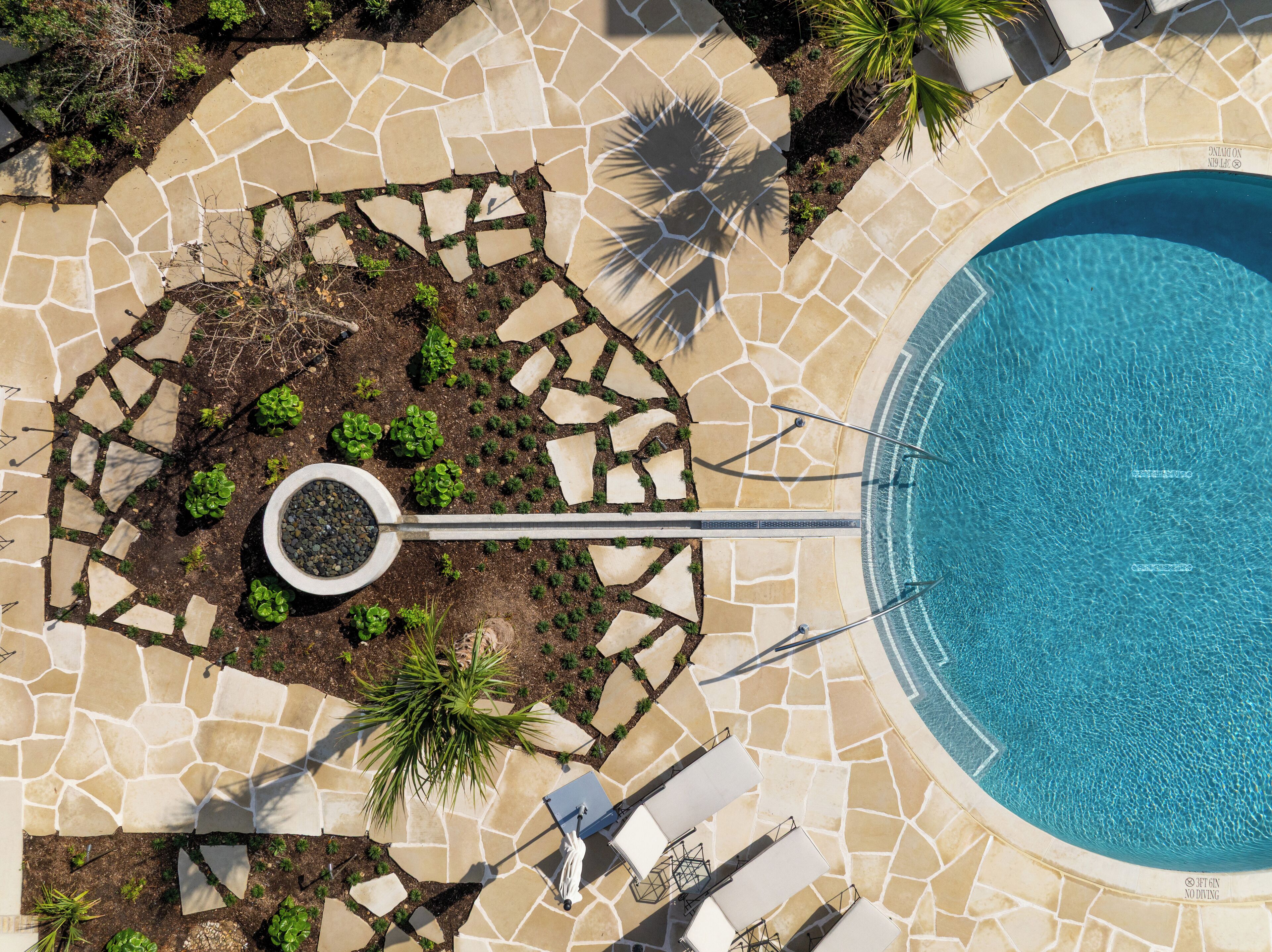 An aerial view of a pool area with stone tiles, a circular garden with plants, and lounge chairs.