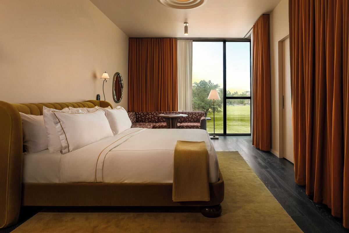 A hotel room with a large bed, a sofa, a small table, and a view of the outdoors through a sliding door.