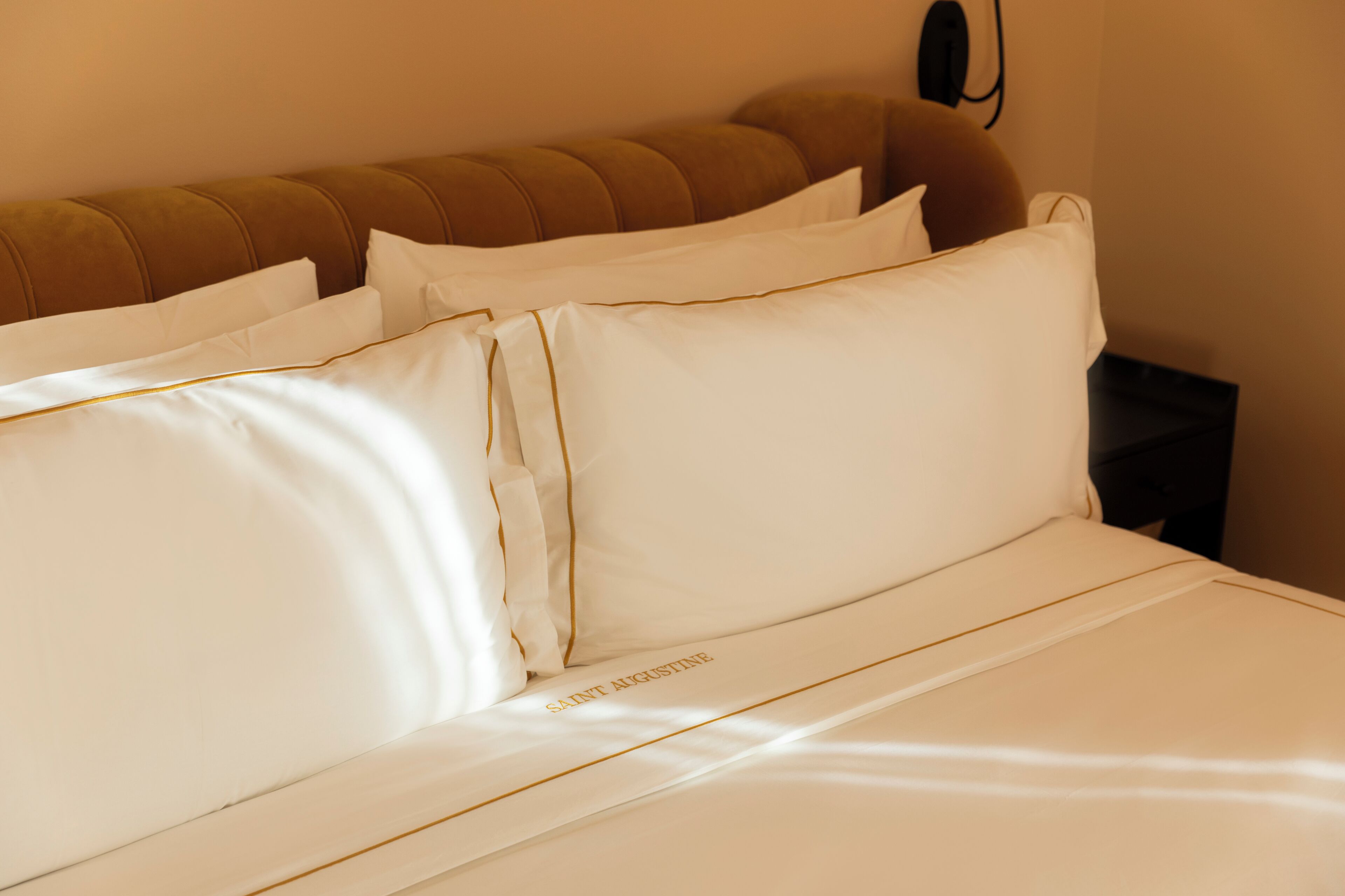A neatly made bed with white linens and gold trim, a brown upholstered headboard, and a black nightstand with a lamp.