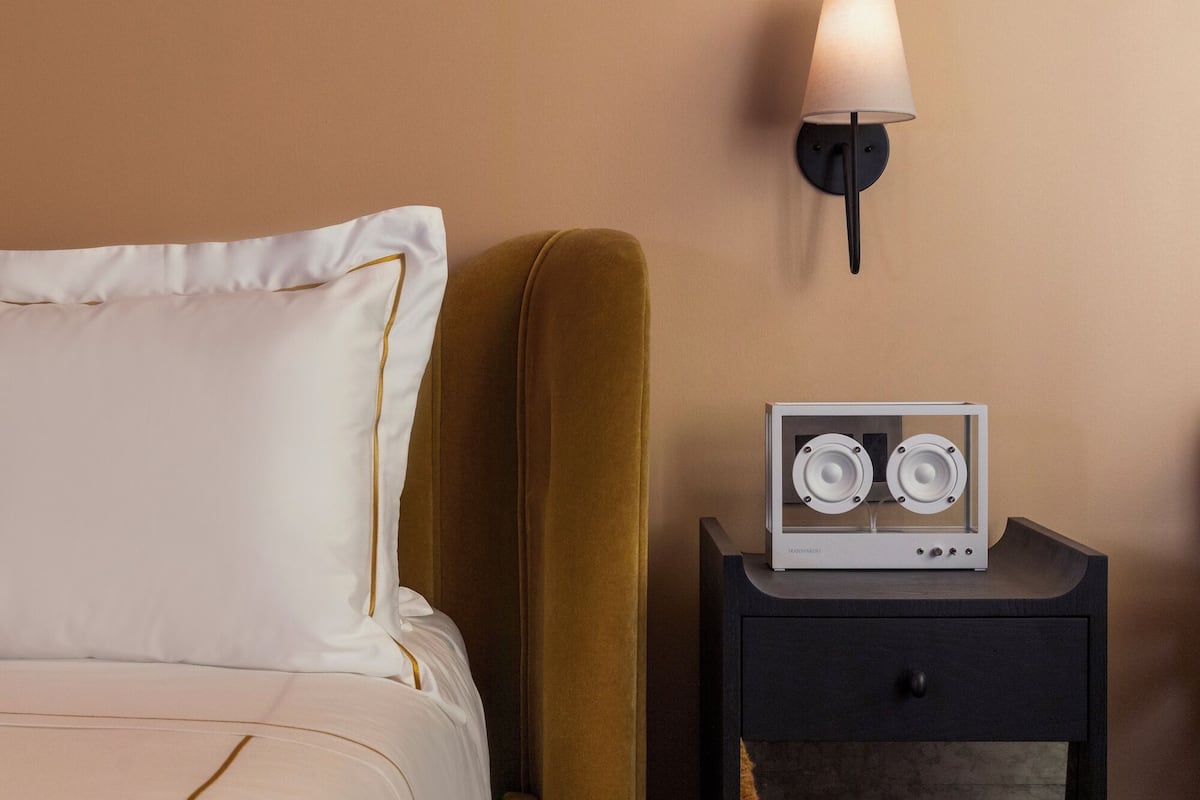 A neatly made bed with a white pillow and a mustard-colored blanket. A small black nightstand with a lamp and a speaker is beside the bed.