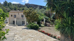 A stone-paved courtyard with a building, lush greenery, and a mountainous backdrop. - Masia Bernat: Antigua Masía en la Naturaleza Para Grupos (Tortosa)
