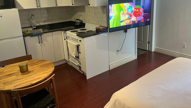 Eine kleine Küche mit weißen Schränken, einem Herd und einer Mikrowelle. Ein Flachbildfernseher an der Wand zeigt einen farbenfrohen Zeichentrickfilm.