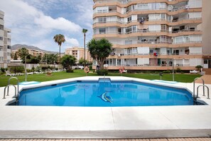 Una piscina con diseño de delfín, rodeada por una terraza blanca, y un edificio de varios pisos al fondo.