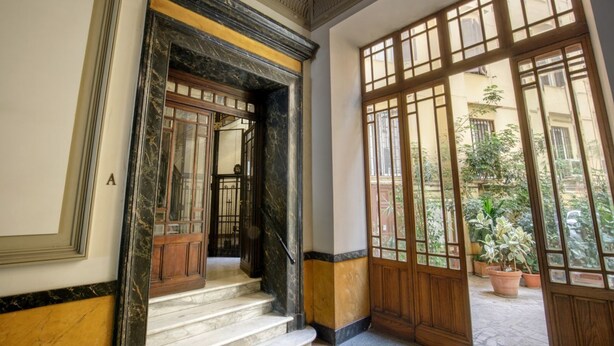 A grand entrance with marble steps and a wooden door, leading to a courtyard with potted plants.