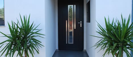 Una entrada de edificio moderno con una puerta negra, dos plantas en macetas a cada lado y unos pocos escalones que conducen a la puerta.