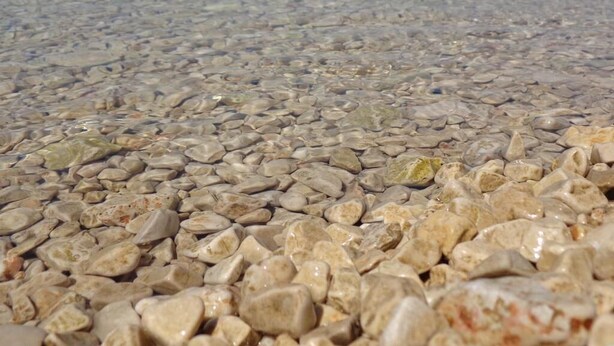 Een kiezelstrand met helder water en bergketens in de verte.