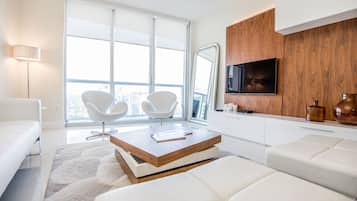 A modern living room with white furniture, a wooden coffee table, a flat-screen TV, and a large window with a city view.