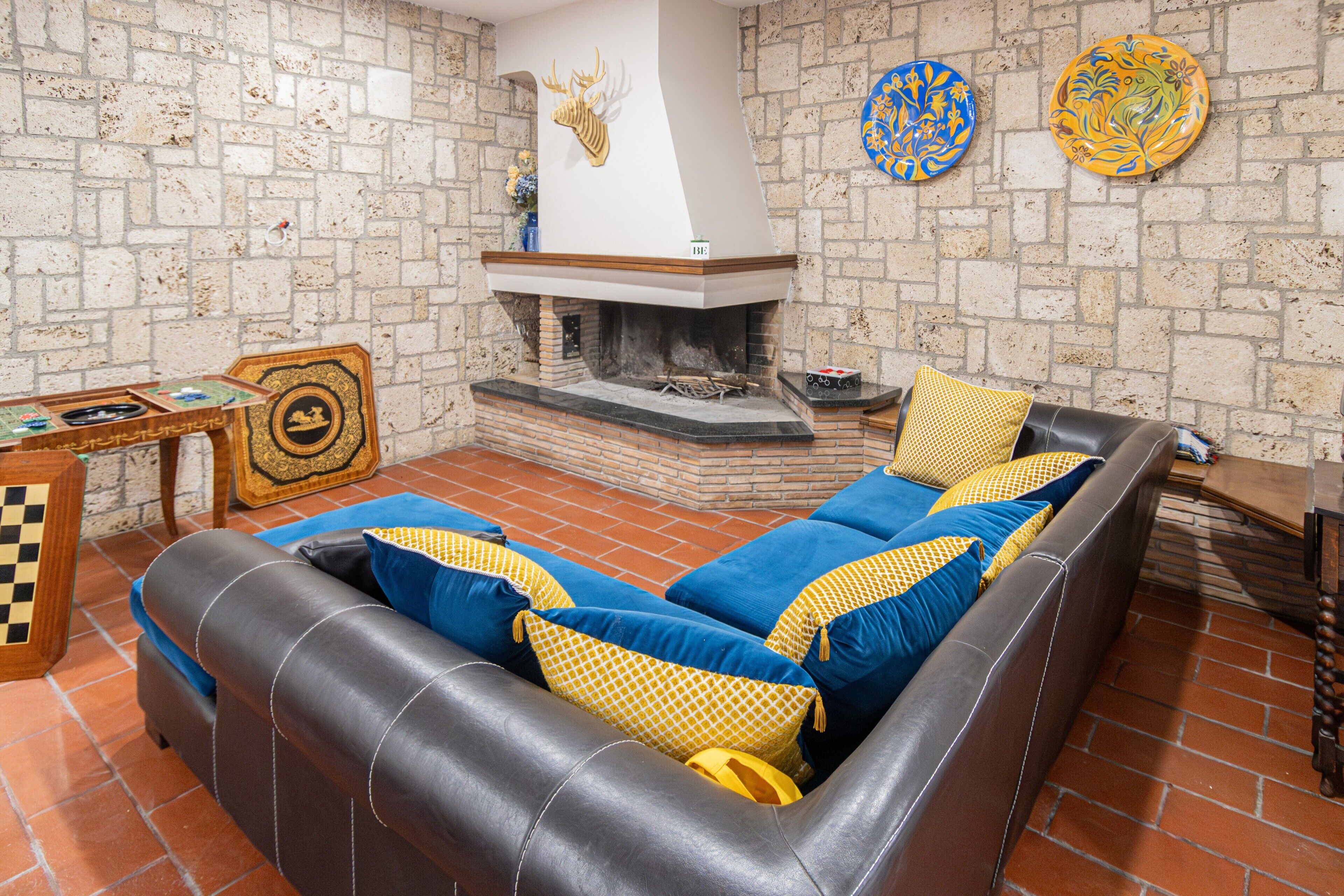A living room with a fireplace, a sofa with patterned cushions, a chessboard, and decorative plates on the wall.