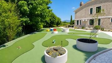 Un campo da minigolf con superficie verde, fioriere bianche con piante e un edificio con finestre sullo sfondo.