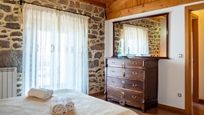 A room with a stone wall, wooden ceiling, a bed with rolled towels, a dresser with drawers, and a mirror. - Country House 'Casa Rural Del Tormes' with Mountain View, Private Terrace and Private Garden (Puente del Congosto)