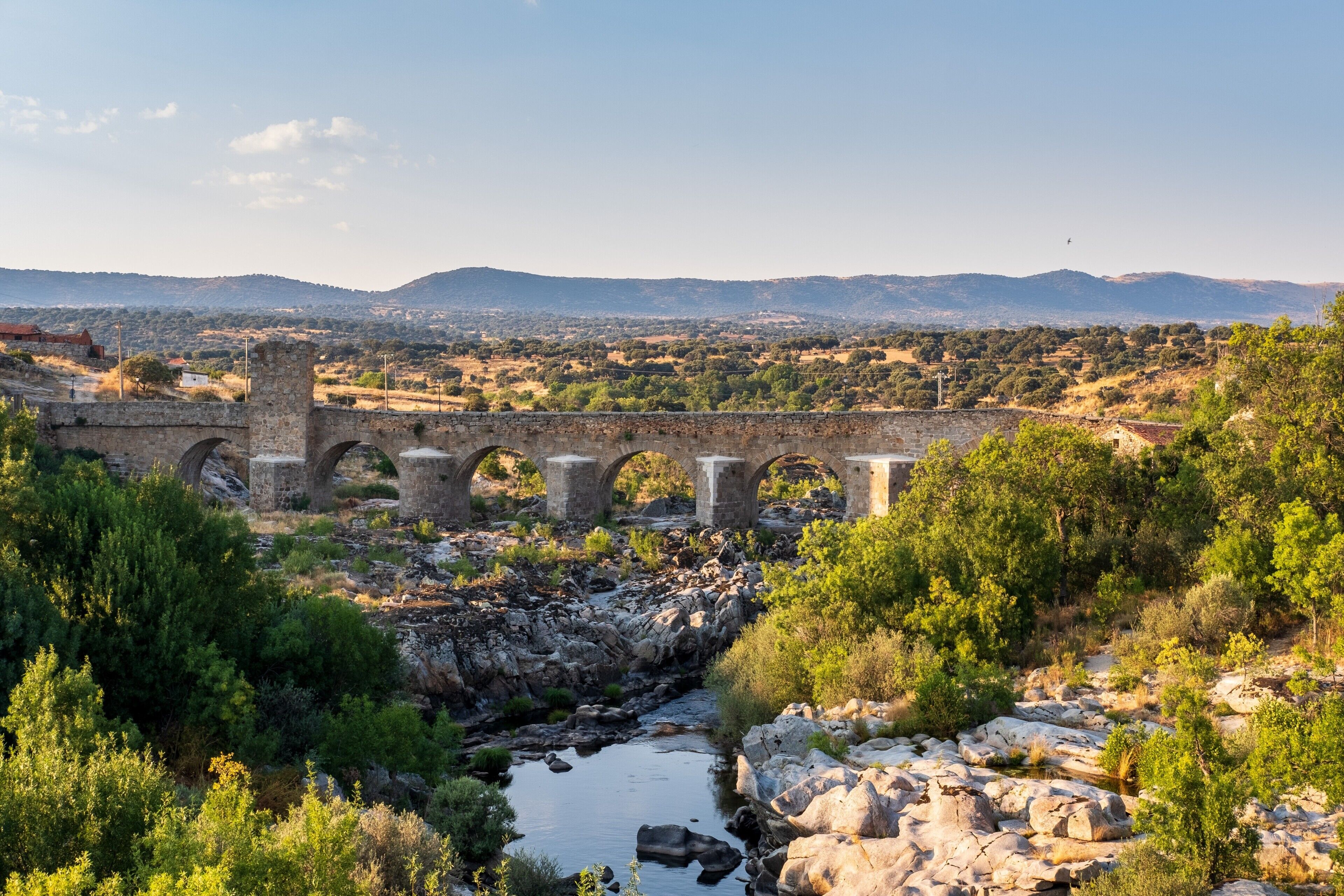 A stone bridge over a river with surrounding greenery and distant hills.