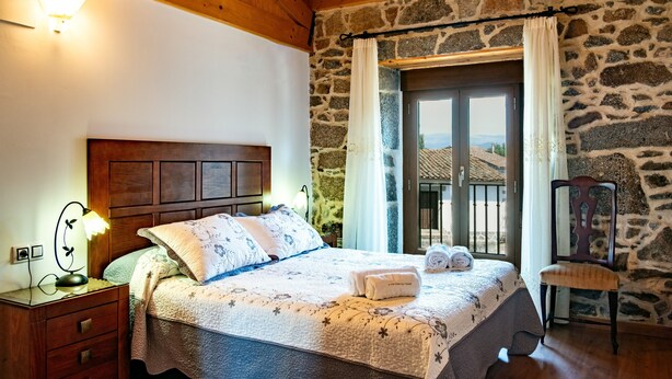 A bedroom with a wooden headboard, a stone wall, a bed with floral bedding, a nightstand with a lamp, and a view through a window. - Country House 'Casa Rural Del Tormes' with Mountain View, Private Terrace and Private Garden (Puente del Congosto)