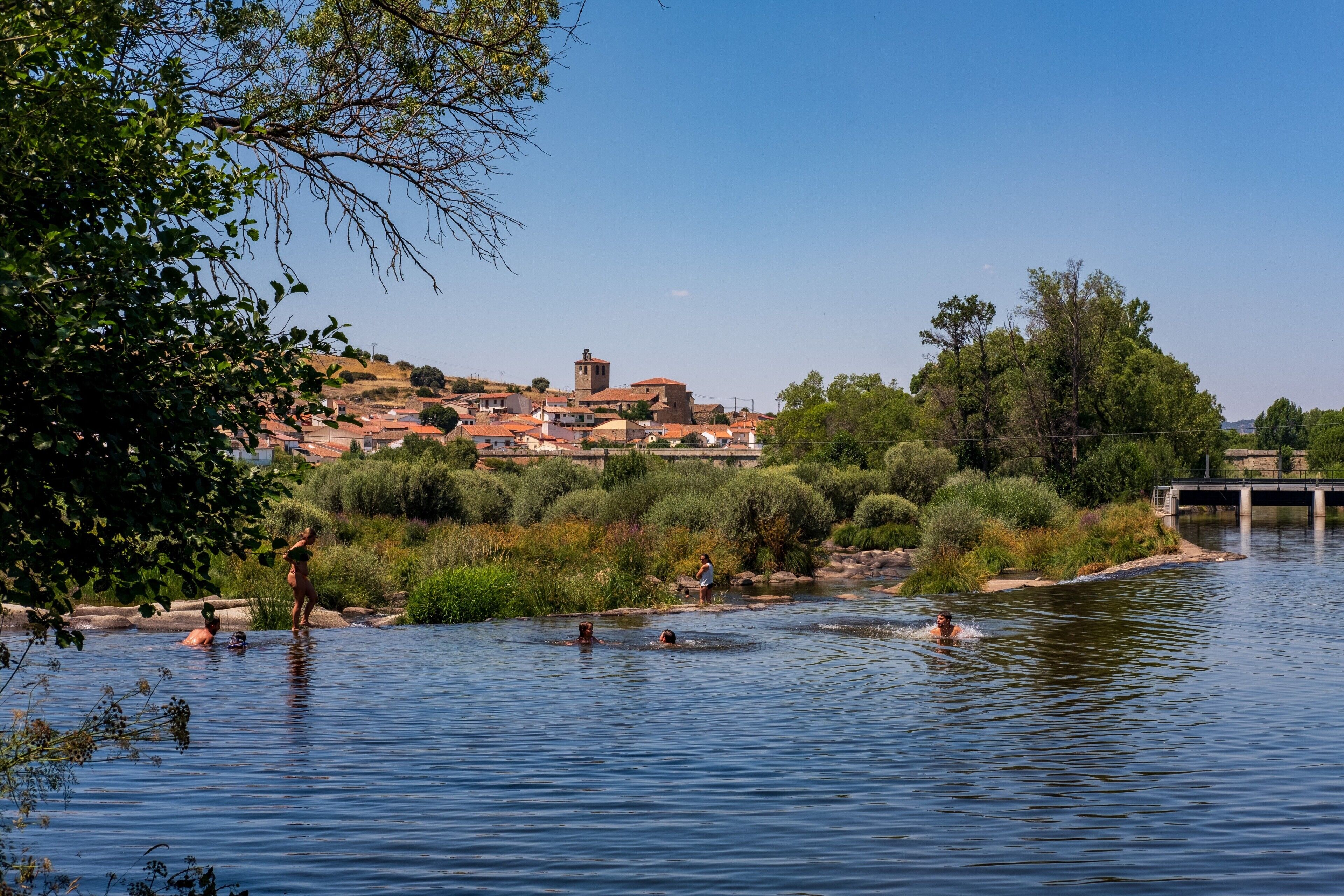 Une rivière où des gens se baignent, un pont et une ville avec des bâtiments en arrière-plan.