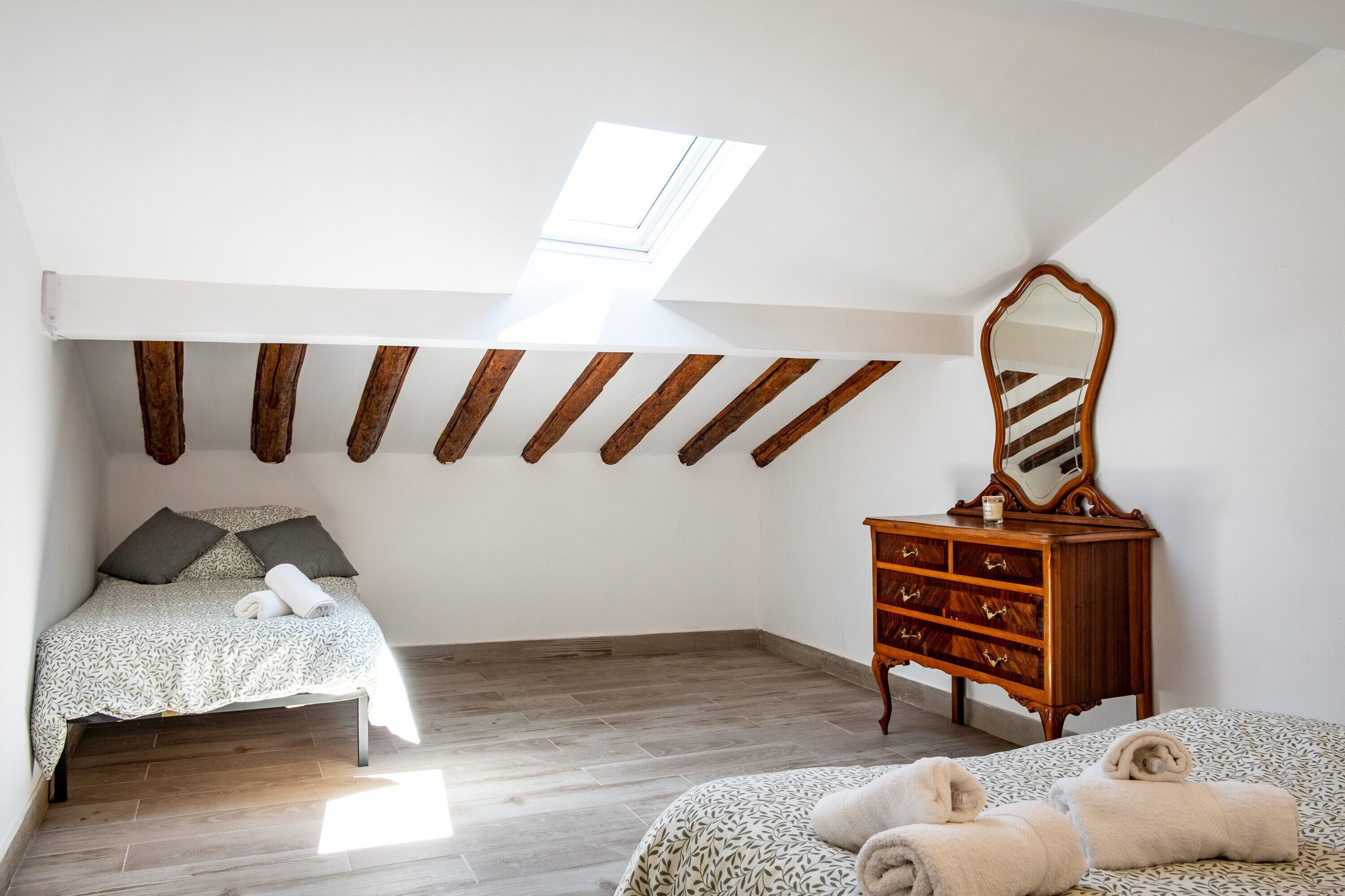 Un dormitorio con techo inclinado, vigas de madera, cama con colcha estampada, cómoda con espejo y tragaluz.