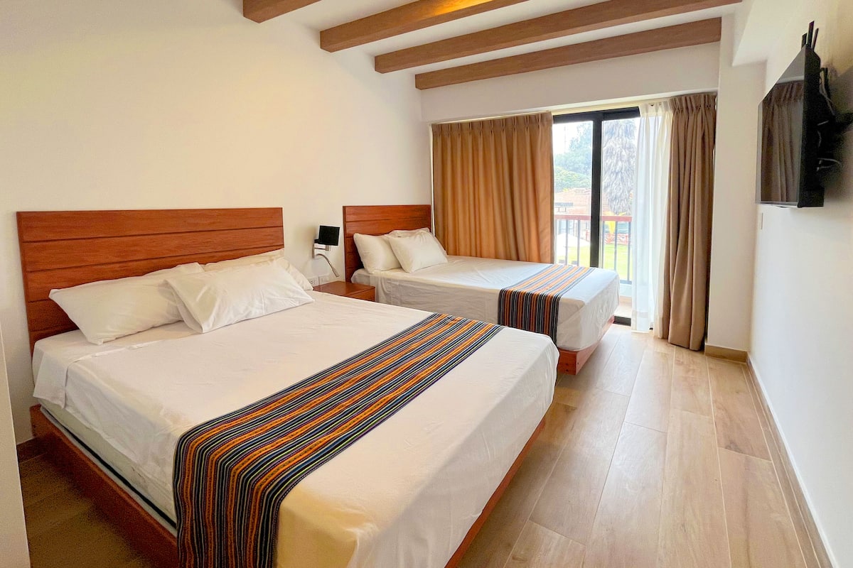 A hotel room with two beds, wooden headboards, a large window with curtains, and a flat-screen TV mounted on the wall.