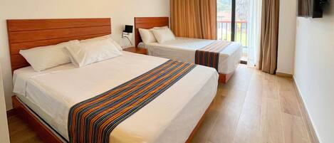 A hotel room with two beds, wooden headboards, a large window with curtains, and a flat-screen TV mounted on the wall.