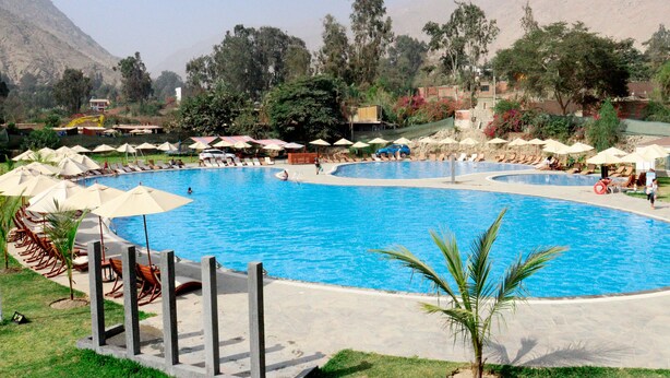 Ein großer, sauberer Pool mit mehreren Liegestühlen und Sonnenschirmen, umgeben von viel Grün und Bergen im Hintergrund.