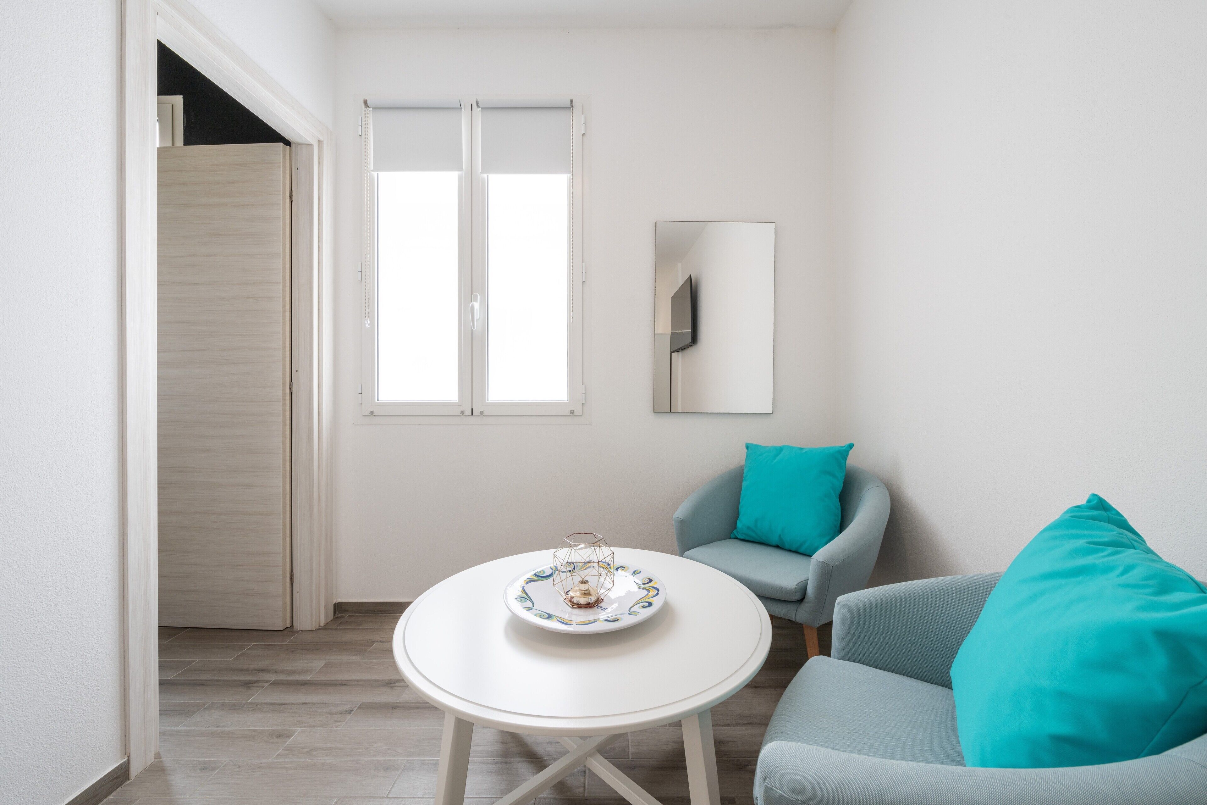 Una stanza con un tavolo bianco, un divano azzurro tenue, cuscini color verde acqua e una porta in legno, oltre a una finestra.