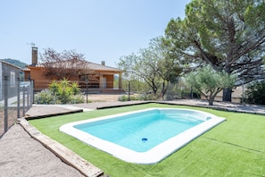 Una piscina rectangular con borde blanco, rodeada de césped artificial verde, una casa con techo de tejas y árboles al fondo.