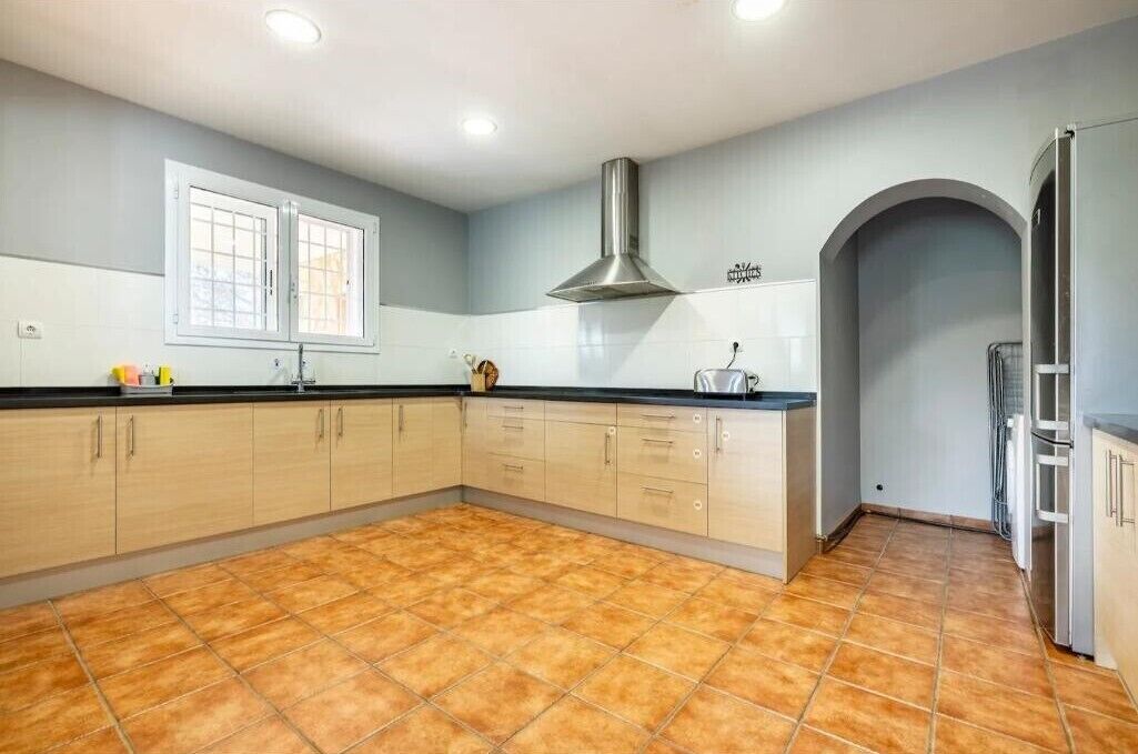 Een keuken met lichte kastjes, een zwart aanrecht, een roestvrijstalen afzuigkap en een raam met witte kozijnen.