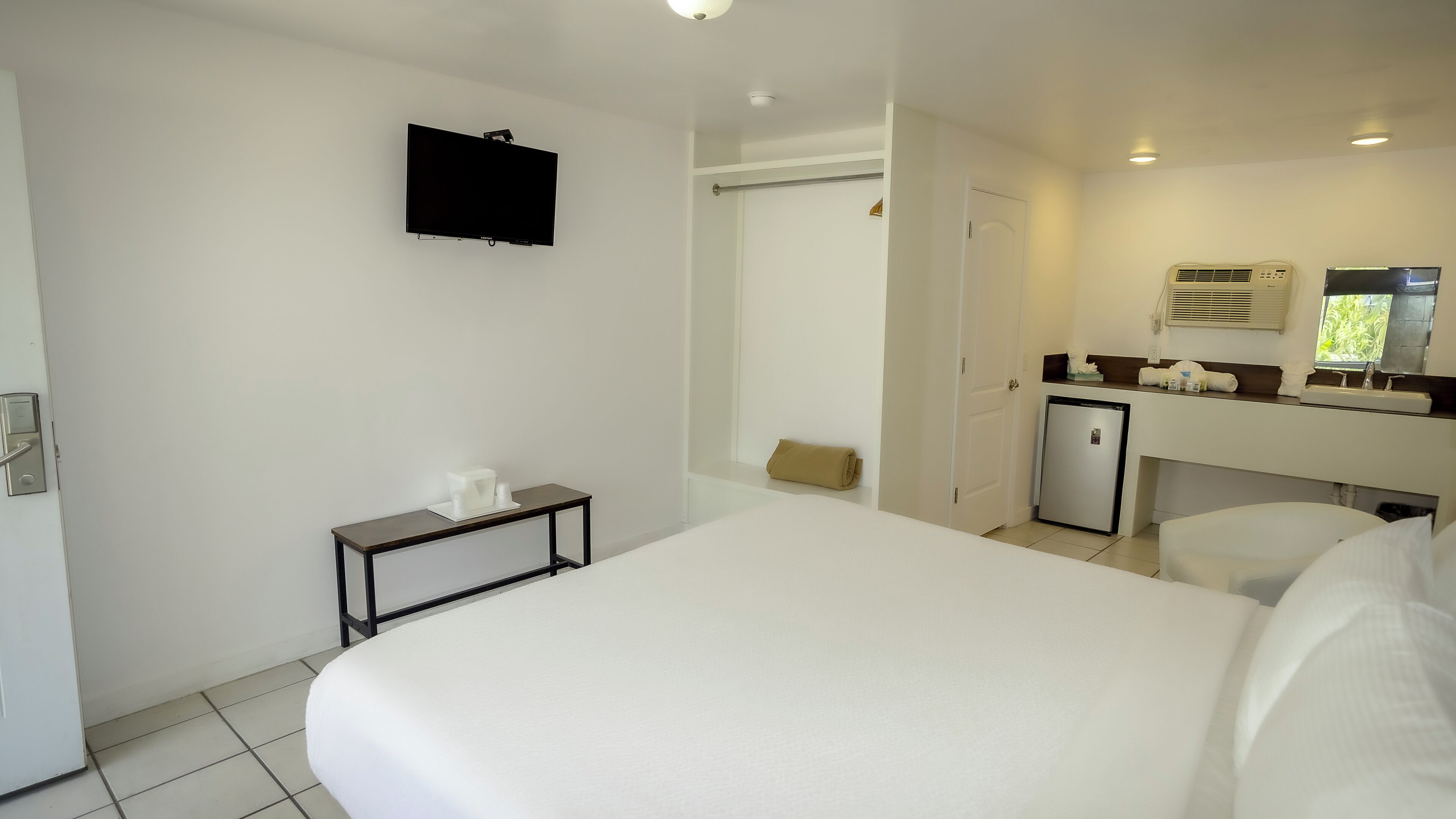 Hotelkamer met een groot bed, een klein nachtkastje met zakdoekendoos, een aan de muur bevestigde televisie, een minibar en een badkamer met spiegel, plus een deur naar een andere kamer.