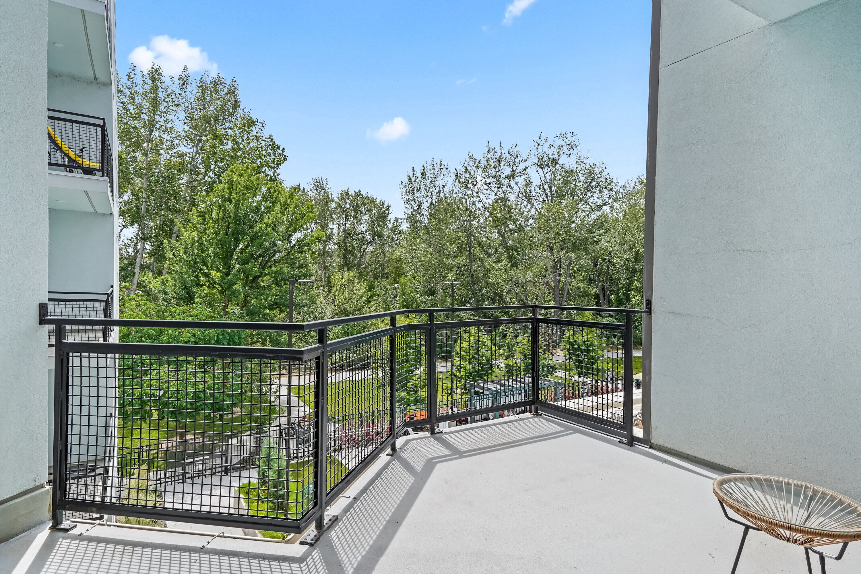 Ein Balkon mit schwarzem Metallgeländer, einem Korbstuhl und Blick auf einen Park mit Bäumen und einem Weg.