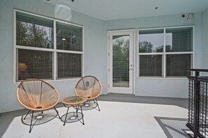 A covered patio area with two wicker chairs and a small table, large windows with blinds, and a door. - Kasa on the River Boise (Boise)