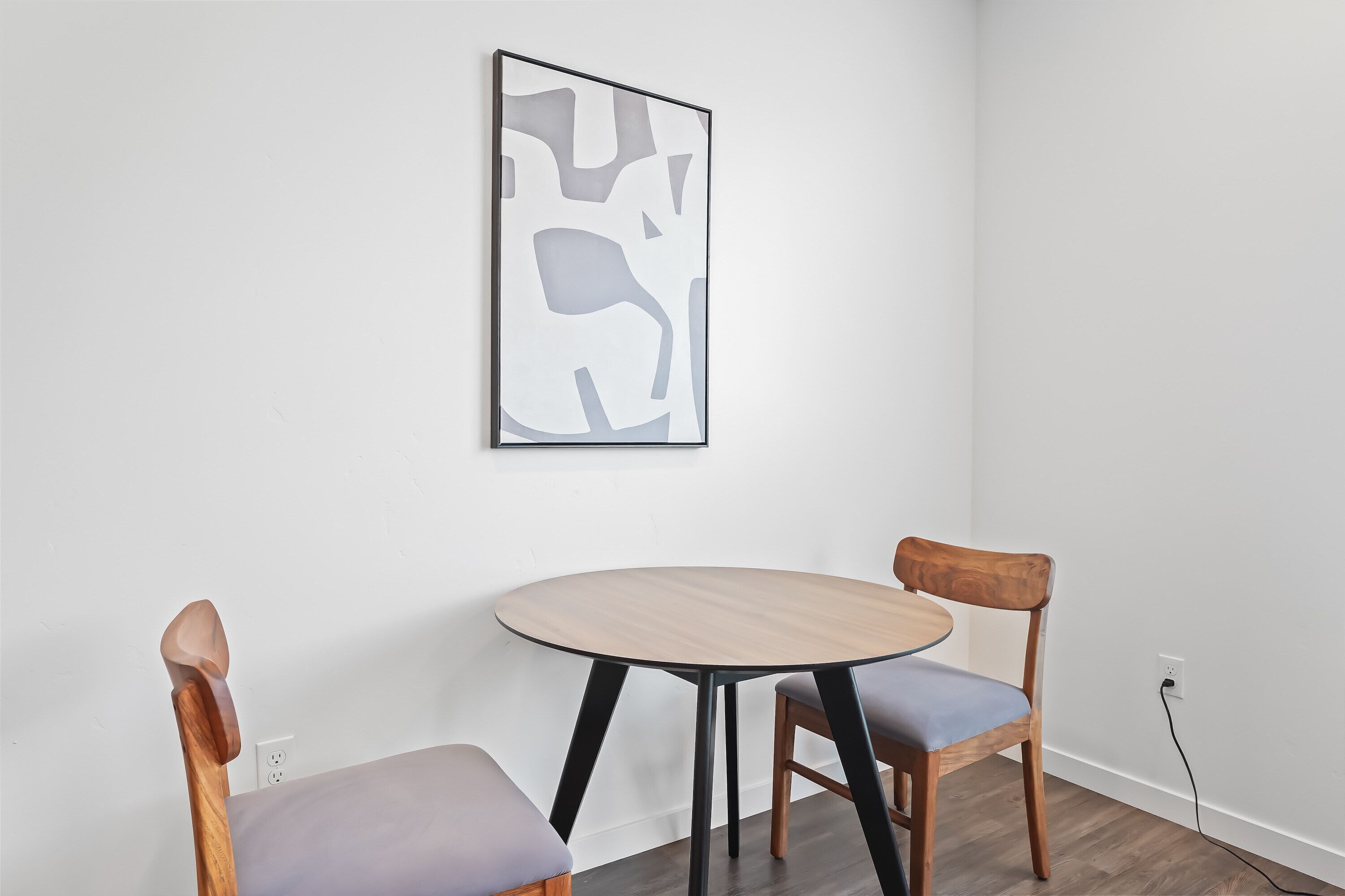 A room with a round wooden table and two chairs, a framed abstract artwork on the wall, and a power outlet on the wall.