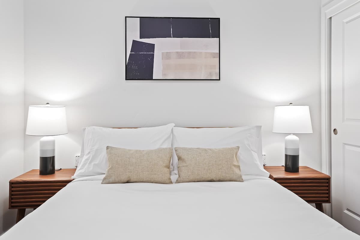 Una habitación de hotel con una cama bien hecha, dos mesitas de noche con lámparas y un cuadro abstracto enmarcado en la pared.