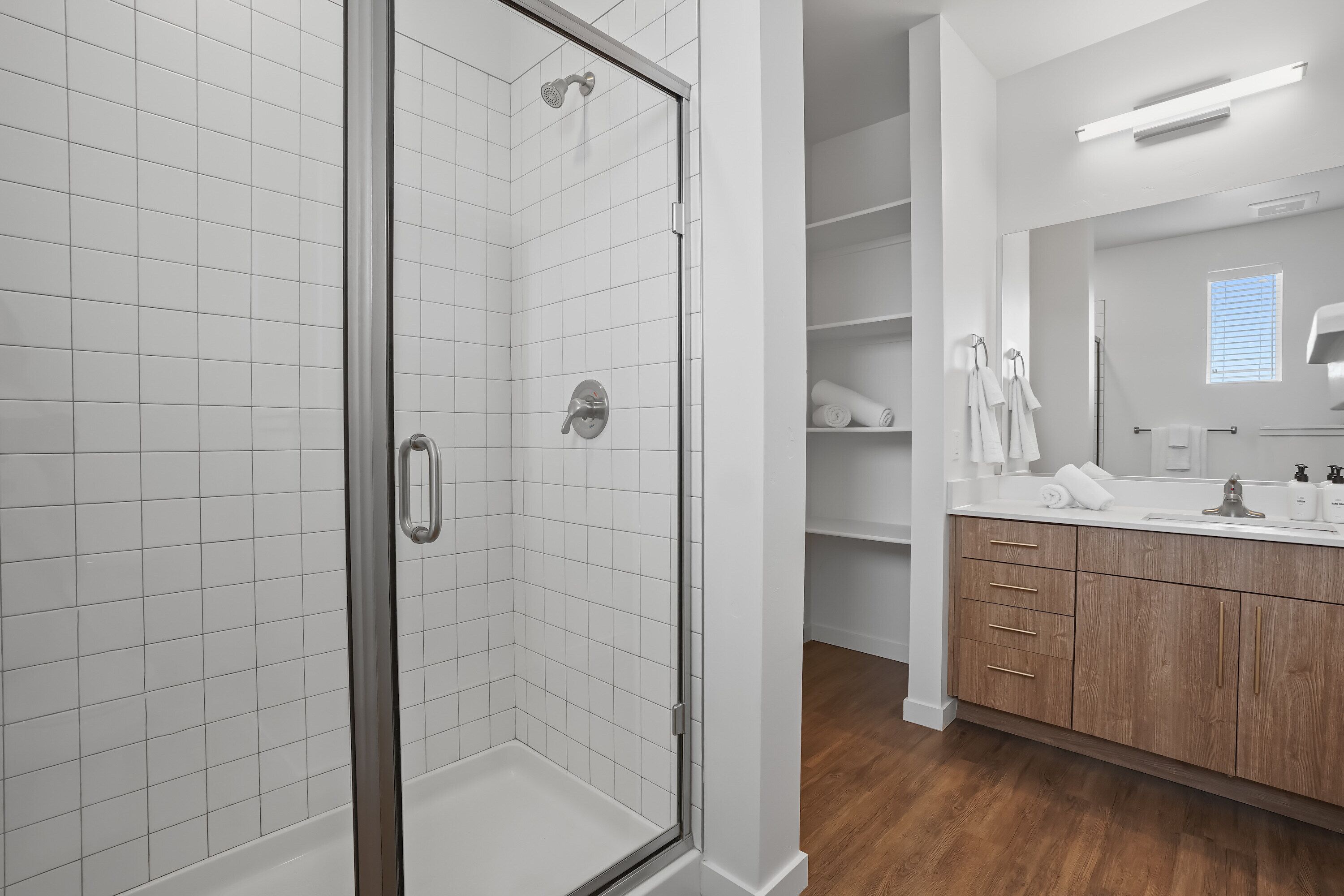 A modern bathroom with a glass-enclosed shower, white tiled walls, wooden cabinets, and a large mirror.