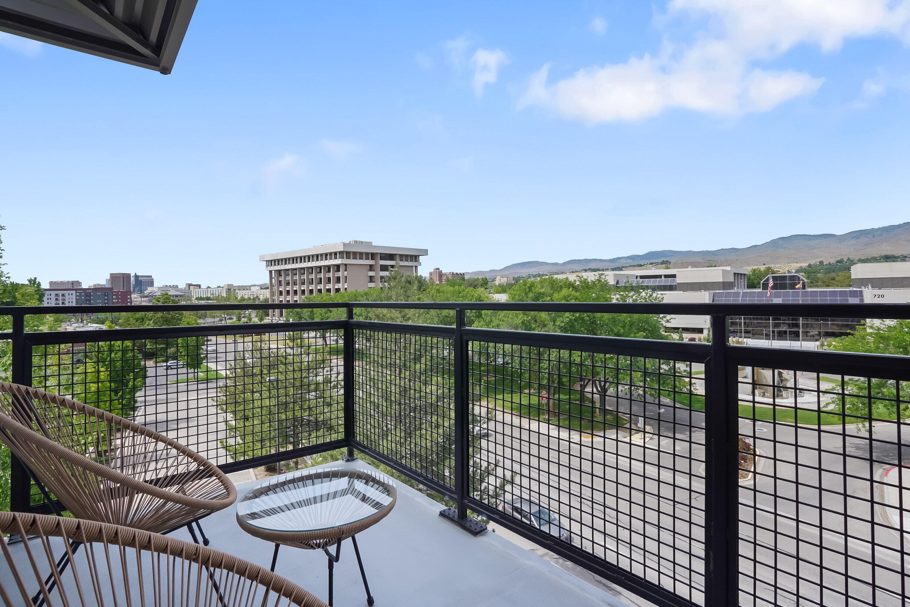 Een balkon met rieten stoelen en een glazen tafel met uitzicht op een stadsgezicht met gebouwen en een weg.