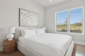 Une chambre à coucher avec un grand lit, une table de chevet avec une lampe, un tableau décoratif au mur et une fenêtre avec des stores.